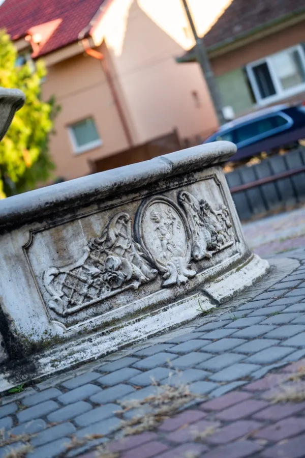 Reliéf na fontáne, za ňou dlažba a dom